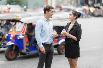 Two business colleagues in street of Bangkok city talking to each other while holding coffee cups.