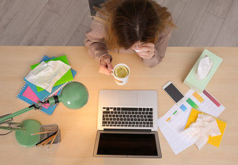 Exhausted woman with tissue and cup of hot drink suffering from cold while working with laptop at table, top view