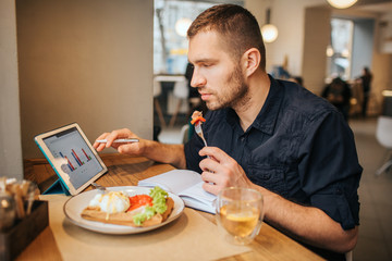 Businessman is sitting at table and eating meal from plate. He has a piece of tomato on fork. Guy is looking at tablet with graphics on its screen. He is wokring.