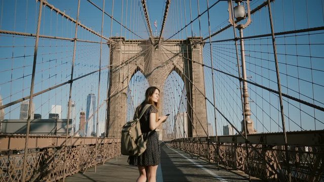 Young European female tourist with backpack walking along Brooklyn Bridge, New York and looking at smartphone 4K.