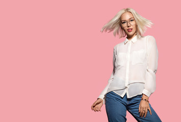 Young attractive model in motion, studio portrait. Stylish hairstyle, fashion glasses, white shirt and jeans. Beautiful blonde lady posing in studio on pink background. Concept of fashion and beauty.