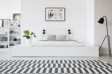 Real photo of a black lamp standing next to a white platform with a bed and a monstera plant on it in modern hotel bedroom interior