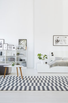 White interior of a cozy bedroom with gray bed on a platform and a wooden table on a striped rug. Real photo