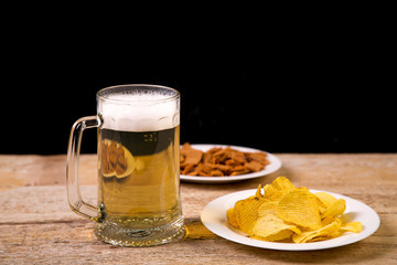 On a wooden table there are plates with crispy croutons and potato chips. Near a large mug with beer. Horizontal image.
