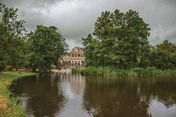 Fototapeta premium Tranquil lake with lawn, grove. building and cloudy sky in Amsterdam park. Famous for its huge cultural activity, graceful canals and bridges. Northern Netherlands.