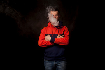 A young dark-haired man in a colored orange and blue hoody standing in close posture smokes a vape and exhales a white smoke on a black isolated background