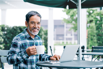Portrait of happy white stylish short beard mature man drinking coffee while using laptop. Casual...