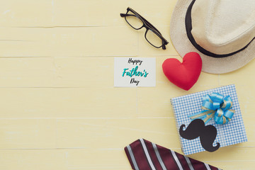 Happy fathers day concept. Red tie, glasses, hat, mustache, gift box with Happy father's day text and handmade red heart on bright yellow pastel wooden table background.