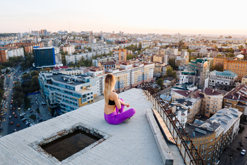 Young beautiful girl in sports uniform sits on the edge of the roof in a lotus pose during sunset and looks into the horizon.