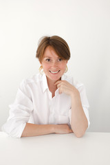 Naklejka premium Beautiful adult woman in white shirt sitting at desk on white and smiling at camera