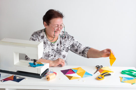 profession, manufacturing, quilting concept. there is old smiling woman in white shirt with floral pattern and glasses, she is sitting at the desk with sewing machine and collecting patches together