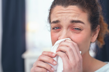 Catching disease. Young tired looking girl having her eyes red while cleansing her congested nose suffering from flu