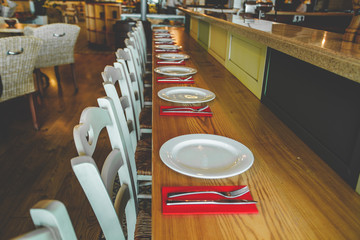 Close-up view of the row of the chairs and table with the white plates and the flatware on the red serviettes. Contemporary interior of the restaurant. Modern decoration in bright colors.