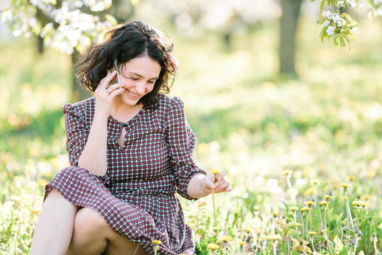connection, beauty, fashion concept. on the grass surrounded by lots of dandelions there is a marvelous woman with dark hair and pale complexion, her radiant smile is illuminating everything