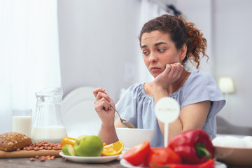 Lack of appetite. Young woman sitting at a breakfast table holding a spoon and looking disheartened while trying to force herself to eat - obrazy, fototapety, plakaty