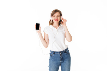 Fototapeta premium Portrait of pretty smiling lady in headphones standing with cellphone in hand and happily looking in camera on white background isolated