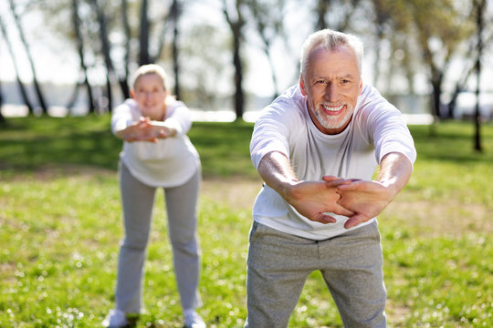 Useful exercise. Cheerful aged man leaning forward while doing exercises