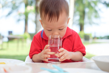 Cute little Asian 18 months / 1 year old toddler boy child sitting in high chair holding & drinking glass of water by himself at restaurant in beach resort, best beverages for child's health concept