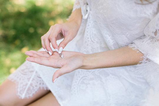 present, jewel, accessory concept. fair-skinned woman wearing white dress with laces negligee taking two beautiful dizzling silver ear-rings with clear transparent diamonds shining in the light of sun