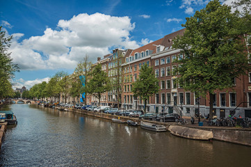 Tree-lined canal with old brick buildings, steeple, moored boats and sunny blue sky in Amsterdam. The city is famous for its huge cultural activity, graceful canals and bridges. Northern Netherlands.