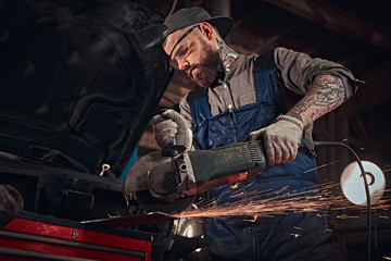 Brutal auto mechanic in a uniform and safety glasses working with an angle grinder while standing...