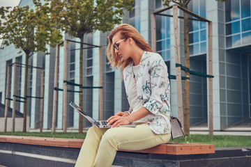 Stylish fashion blogger using the laptop for work while sitting outside on a bench against a skyscraper.