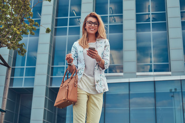 Fashionable female blogger in stylish clothes and glasses with a handbag, holds a smartphone, standing against a skyscraper.