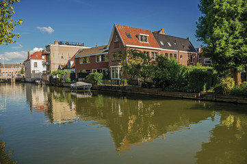 Fototapeta na wymiar Wide canal with brick houses and boat moored on its bank reflected in water under blue sky of sunset in Weesp. Quiet and pleasant village full of canals and green near Amsterdam. Northern Netherlands.