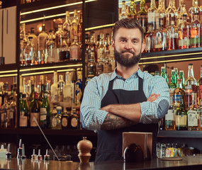 Stylish brutal bartender in a shirt and apron standing with crossed arms at bar counter background.