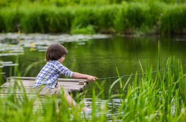 A boy in a plaid shirt sitting on a wooden bridge on a overgrown river and fishing branch