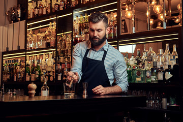 Stylish brutal barman in a shirt and apron makes a cocktail at bar counter background.