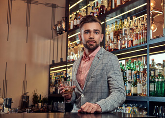 Stylish brutal barman in a shirt and apron makes a cocktail at bar counter background.