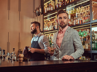 Stylish brutal barman in a shirt and apron makes a cocktail at bar counter background.