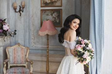 Beautiful fashionable bride, young model brunette woman in stylish wedding dress with naked shoulders with bouquet of flowers in her hands posing in luxury vintage interior near the window