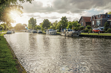 Obraz premium Wide tree-lined canal with houses and boats and shine of sunset reflected in water at Weesp. Quiet and pleasant village full of canals and green near Amsterdam. Northern Netherlands. Retouched photo.
