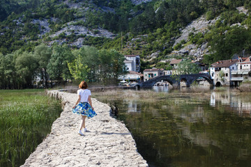 Fototapeta na wymiar girl in a blue skirt going on the asphalt road near the river, green mountains, stone old houses Montenegro