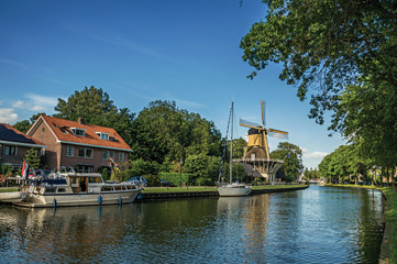 Fototapeta premium Boats and wooden yellow windmill next to wide tree-lined canal on the bright light of sunset at Weesp. Quiet and pleasant village full of canals and green near Amsterdam. Northern Netherlands.