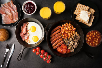 Full English breakfast for two: fried eggs with bacon, mushrooms, beans, grilled sausages in cast-iron frying pans, jam, toast and orange juice on a black stone background. Flat lay. Top view.