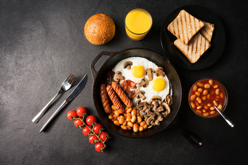 English breakfast in pan with fried eggs, sausages, bacon, beans, tomatoes, bun, toasts and orange juice on dark stone background. Top view. Flat lay.