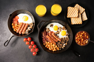 Full English Breakfast for two in cast-iron frying pans with fried eggs, bacon, mushrooms, beans, grilled sausages, tomatoes, toast and orange juice on black stone background. View from above.