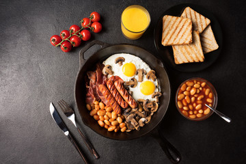 Full English breakfast with fried eggs, sausages, bacon, mushrooms, beans in cast-iron frying pan with toasts and orange juice on copy space black stone background. Flat lay. Top view.