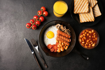 Full English breakfast with fried egg, sausages, tomatoes, beans in cast-iron frying pan with toasts and orange juice on copy space black stone background. Flat lay. Top view.