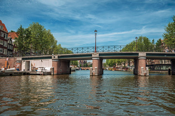 Bridge with iron balustrade on tree-lined canal, old buildings and sunny blue sky in Amsterdam. The...