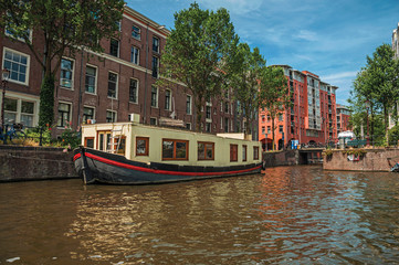Fototapeta na wymiar Big boat used as house moored at side of tree-lined canal with old buildings and sunny blue sky in Amsterdam. Famous for its huge cultural activity, graceful canals and bridges. Northern Netherlands.