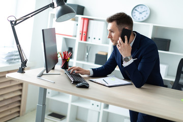 Naklejka premium A young man is standing near a table in the office, talking on the phone and typing text on the keyboard.