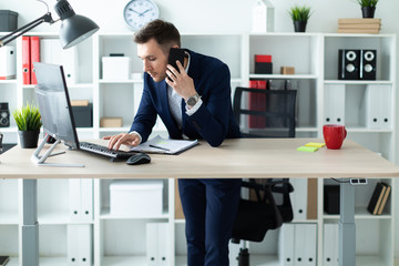 A young man is standing near a table in the office, talking on the phone and typing text on the keyboard.