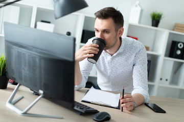 Fototapeta premium A young man stands near a table in the office, holds a pencil in his hand and drinks coffee. A young man works with documents and a computer.
