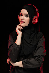 Portrait of beautiful smart young muslim woman wearing black hijab listening to music in headphones on black background