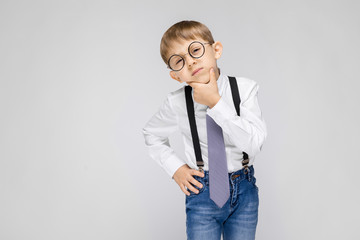 A charming boy in a white shirt, suspenders, a tie and light jeans stands on a gray background. a...