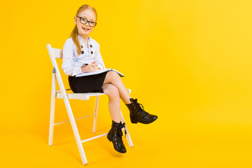 Girl with red hair on a yellow background. A charming girl in transparent glasses sits on a white chair and makes notes in a notebook.